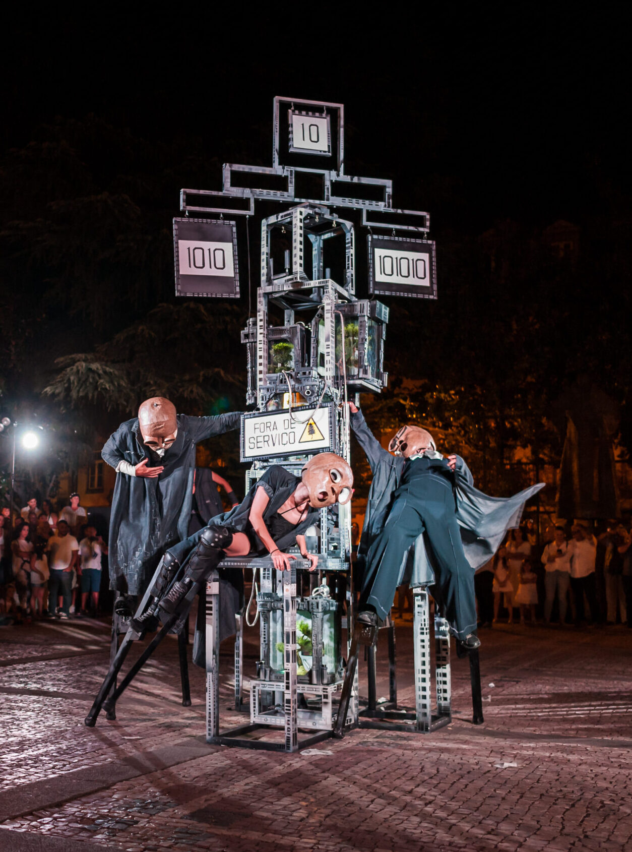 Nighttime street performance featuring performers in dark robes climbing a tall metal sculpture with digital panels displaying 1010, in front of a crowd gathered along a cobblestone square (For a service sign visible).